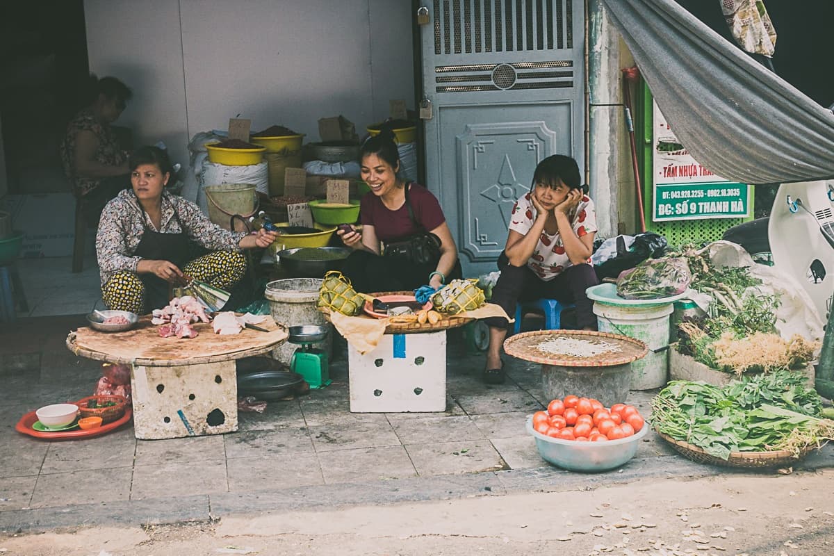 Drie vrouwen zitten bij een kraampje op de straatmarkt met manden vol groenten, kruiden en tomaten. Dit is de levendige sfeer van Streetfood van Azie. De een snijdt, de ander lacht met haar telefoon, terwijl de derde haar kin op haar handen laat rusten.