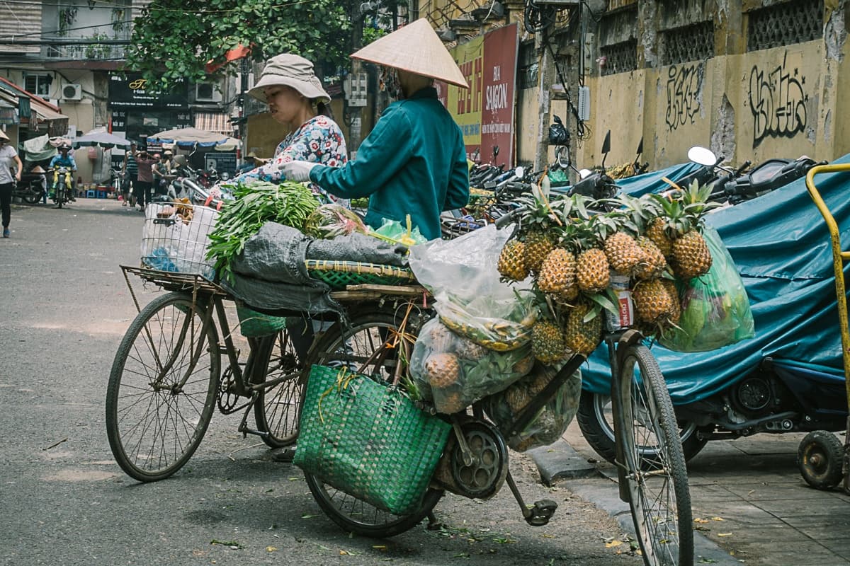 Twee straatverkopers met kegelvormige hoeden staan naast fietsen vol verse producten, die het levendige straatbeeld van streetfood van Azie oproepen, in een drukke stadsstraat met geparkeerde motoren, graffiti en mensen op de achtergrond.