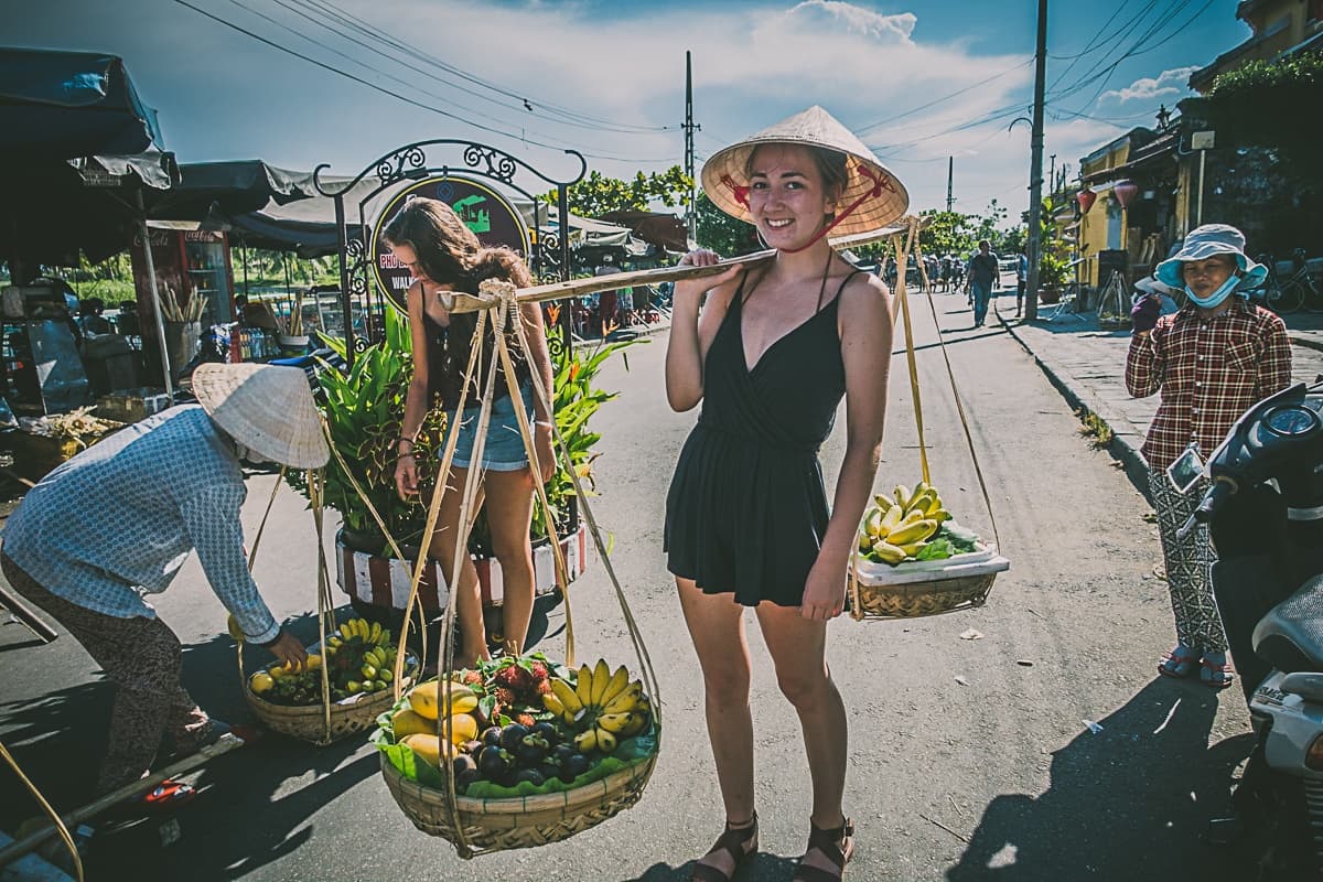 Een vrouw in een zwarte outfit en kegelvormige hoed lacht terwijl ze twee manden met fruit aan een paal draagt op een drukke markt voor streetfood van Azie. Andere mensen, ook met kegelvormige hoeden, schikken fruit in de buurt op de zonnige, drukke straat.