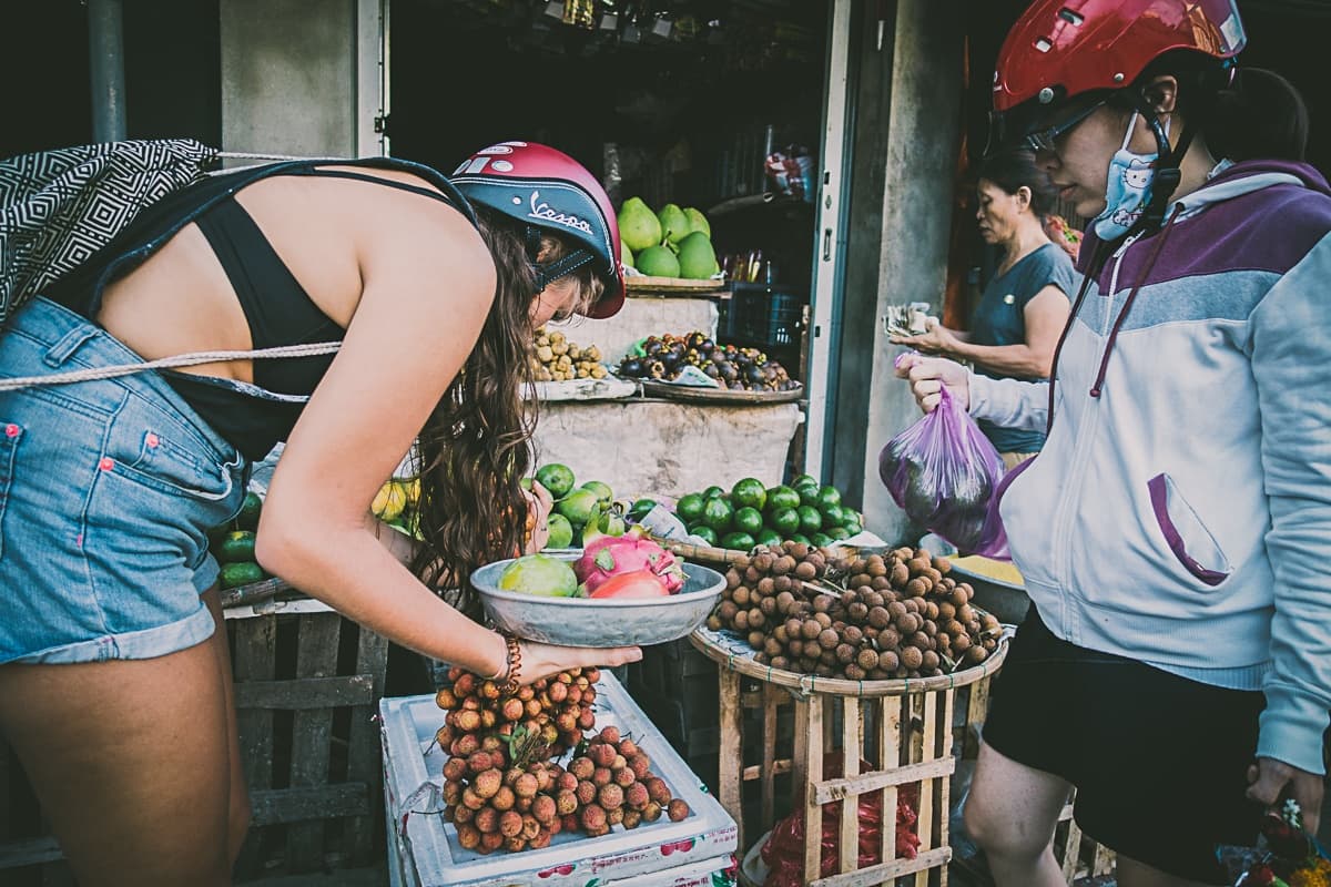 Twee vrouwen met helmen kopen vers fruit bij een kraampje op de markt, dat doet denken aan streetfood van Azie. De ene legt lychees op een weegschaal terwijl de andere een zak vasthoudt. De kraam ligt vol met kleurrijk fruit en op de achtergrond is een andere vrouw te zien.