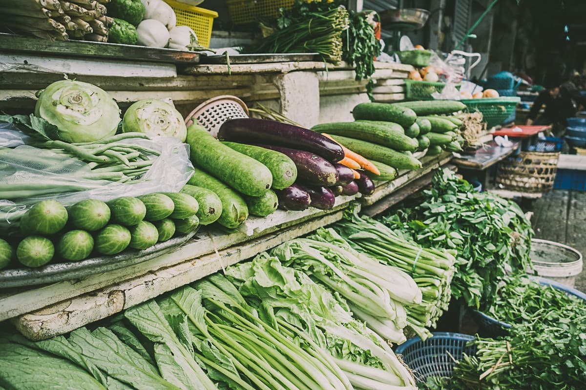 Een marktkraam toont een verscheidenheid aan verse groenten, waaronder komkommers, aubergines, sla, bladgroenten, koolrabi en kouseband - een afspiegeling van het levendige assortiment in streetfood van Azie - netjes gerangschikt op planken en in manden.