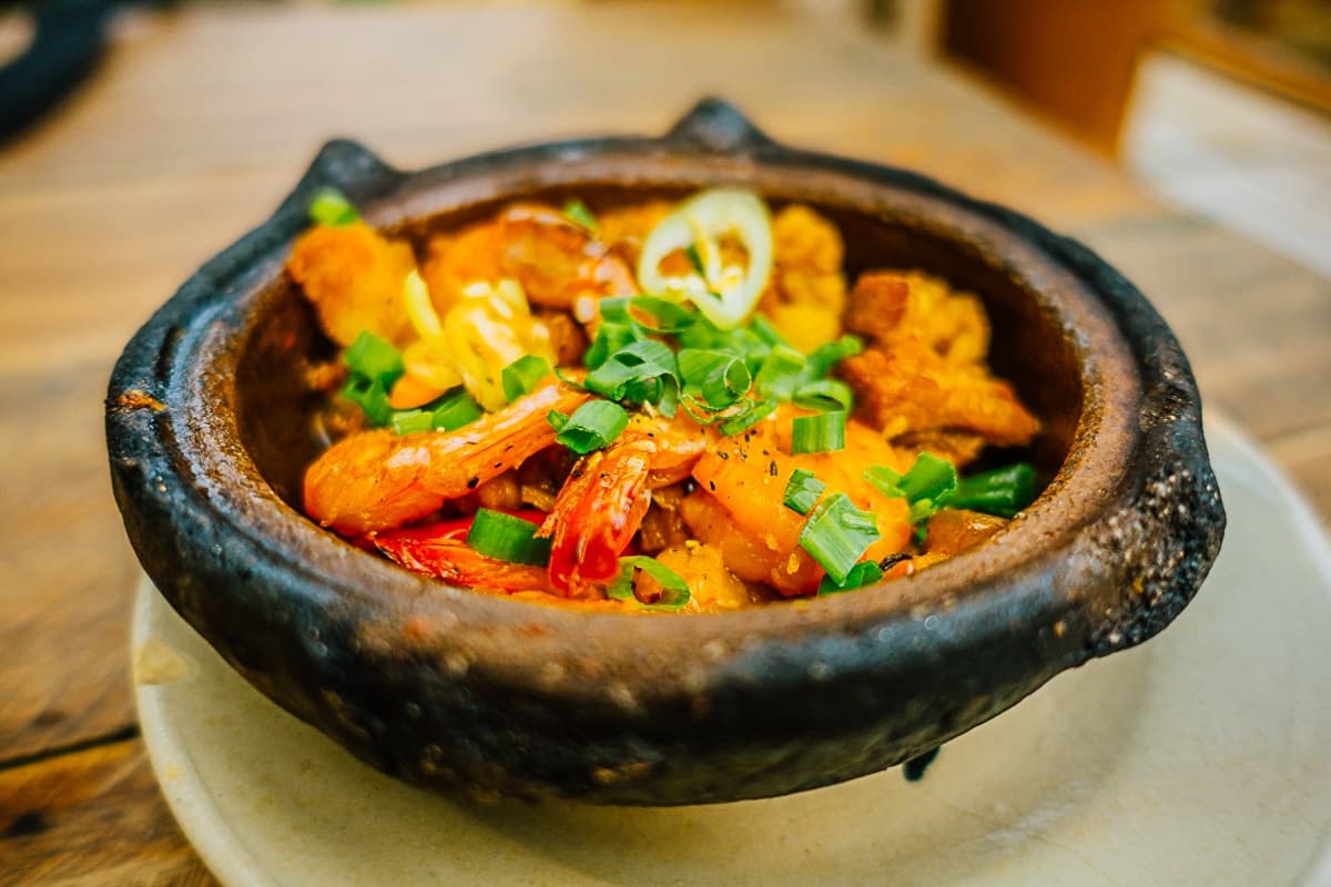 Een rustieke schaal van klei gevuld met een levendig gerecht geïnspireerd door Streetfood van Azie, met garnalen, gehakte groene uien, gele paprika en verse kruiden. Geserveerd op een keramisch bord op een houten tafel ziet de maaltijd er kleurrijk en uitnodigend uit.
