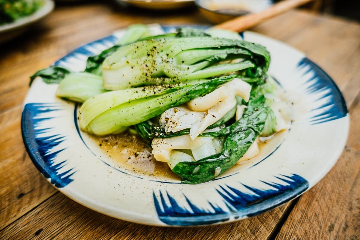 Een close-up van gestoomde bok choy op een decoratief bord, gegarneerd met gebarsten zwarte peper. De glinsterende bladgroenten doen denken aan de verse smaken van streetfood van Azie, met saus die rond de bodem op een houten tafel ligt.