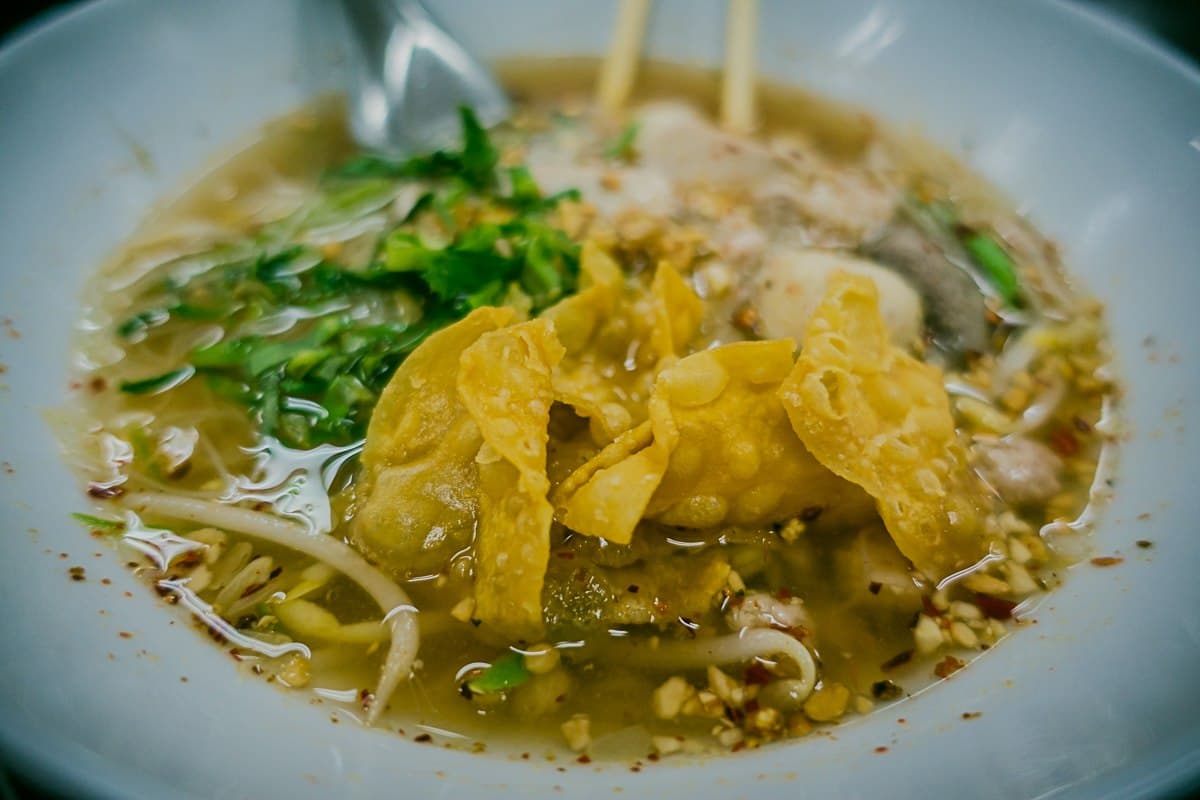 Close-up van een kom noedelsoep, een klassiek streetfood van Azie, met gele gebakken wontons, gehakte groene kruiden, taugé, fijngestampte pinda's en heldere bouillon. Erbij liggen eetstokjes en een metalen soeplepel.