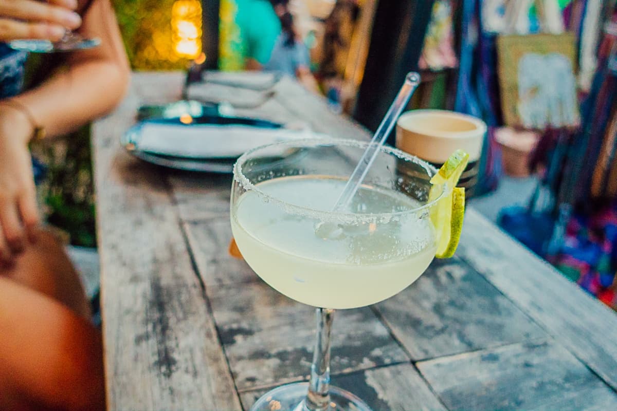 Een margarita-cocktail met een gezouten rand en een garnering van limoendraai ligt op een rustieke houten tafel, die de levendige sfeer van Streetfood van Azie uitstraalt. Op de achtergrond verwijzen borden en een levendig decor naar een dinerervaring in de buitenlucht.