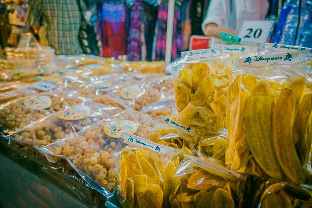 Doorzichtige plastic zakken gevuld met gedroogde bananenchips en andere streetfood van Azie snacks staan uitgestald op een marktkraamtafel. Kleurrijke stoffen hangen op de achtergrond en op een prijsbordje staat "20", waarmee de levendige sfeer van een Aziatische markt wordt weergegeven.