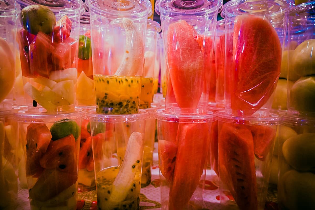Doorzichtige plastic bekertjes gevuld met kleurrijk, gesneden fruit zoals watermeloen, banaan, passievrucht en kiwi staan dicht op elkaar gestapeld, waardoor een levendige display ontstaat die doet denken aan streetfood van Azie op een levendige markt of eetkraam.