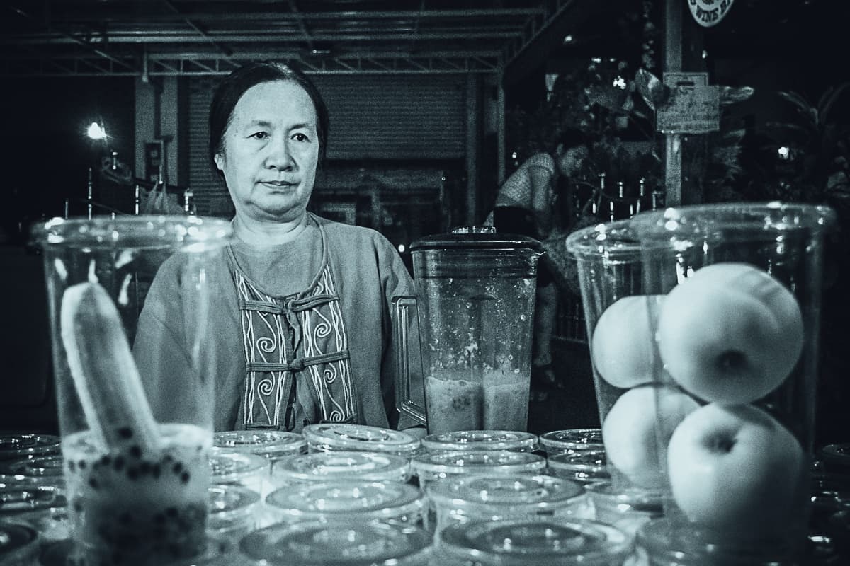 Een vrouw met een neutrale uitdrukking zit achter een tafel bedekt met plastic bekertjes met fruit, zoals appels en een banaan, in wat lijkt op een Aziatische marktkraam met streetfood. De afbeelding heeft een koele blauwe monochrome tint.
