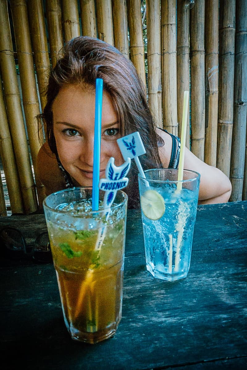 Een vrouw met roodbruin haar glimlacht terwijl ze over een donkere houten tafel leunt. Voor haar staan twee hoge drankjes van streetfood van Azie, de ene gegarneerd met limoen en de andere met munt. Bamboe palen vormen de uitnodigende achtergrond.