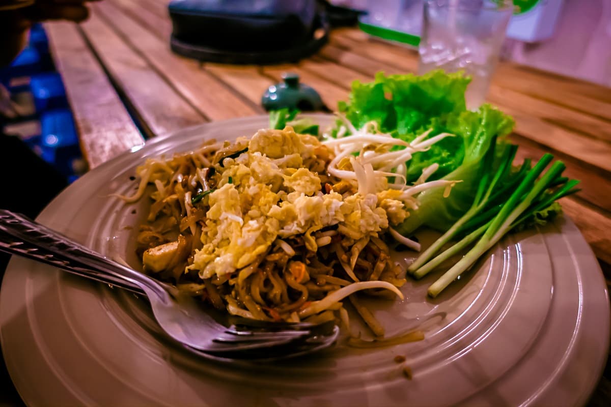 Op een houten tafel staat een bord Pad Thai met roerei, taugé, sla en groene uien - een klassiek gerecht dat je vaak bij streetfood van Azie vindt. Een vork en lepel liggen op het bord, met een glas ijswater op de achtergrond.