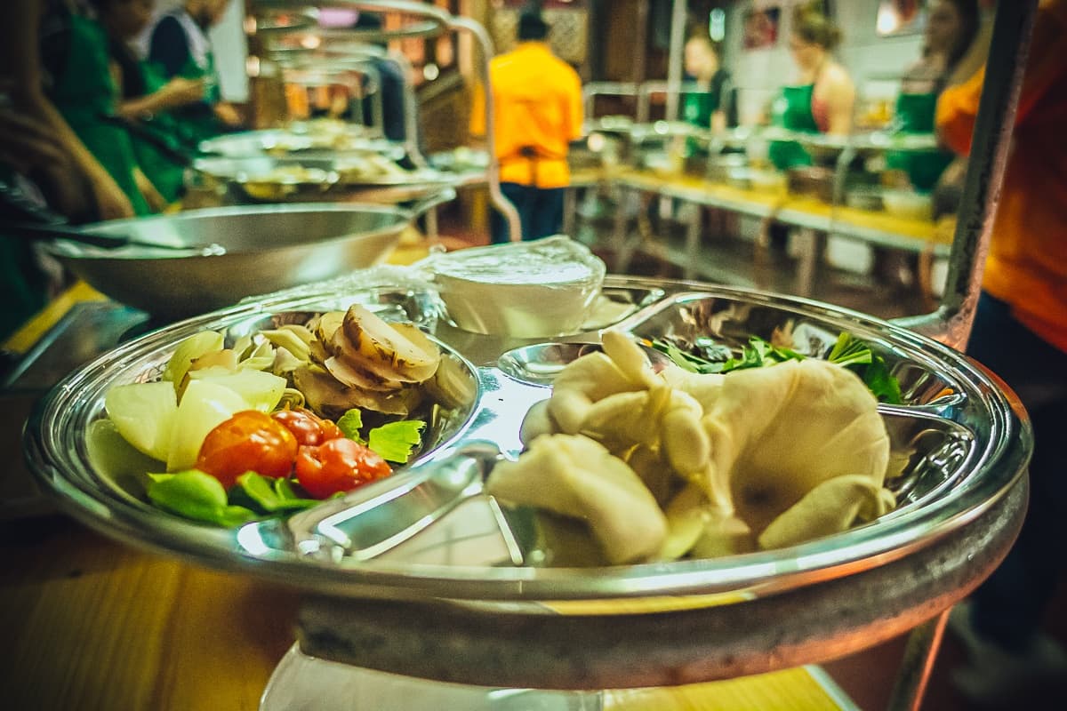 Een metalen cafetaria dienblad met vakjes bevat cherrytomaatjes, sla, champignons en een witte saus. Op de achtergrond staan mensen in groene en oranje shirts in de rij met dienbladen in een drukke kantine, wat het levendige gevoel van streetfood van Azie weergeeft.