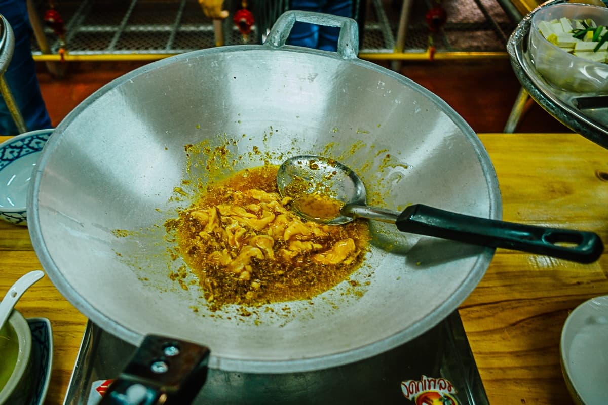Een metalen wok op een fornuis bevat stukken voedsel, waarschijnlijk kip, die worden geroerbakt met bruine kruiden, wat doet denken aan Streetfood van Azie. Een metalen pollepel en een spatel met zwarte steel rusten in de wok temidden van schalen met extra ingrediënten op houten werkbladen.