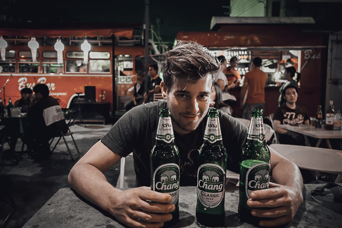 Een man met warrig bruin haar zit 's avonds aan een tafel in de open lucht, lachend achter drie grote groene Chang-bierflessen. Op de achtergrond dineren mensen terwijl rood streetfood van Azie oplicht, wat een levendige, stedelijke sfeer creëert.