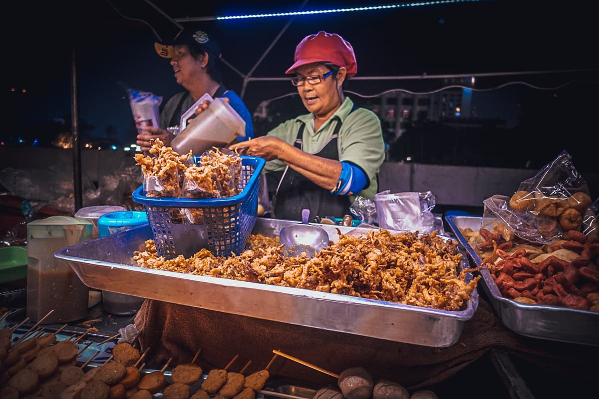Twee straatvoedselverkopers staan bij een kraampje op de avondmarkt, één met een rode hoed en bril, omringd door dienbladen met gefrituurde snacks en spiesjes - een levendig tafereel dat doet denken aan een Streetfood van Azie onder neonverlichting.
