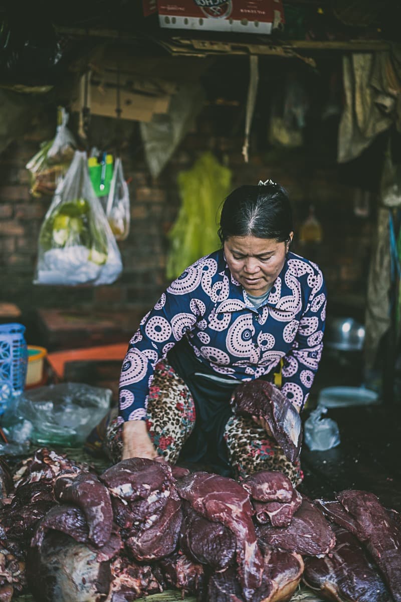 Een vrouw met een gedessineerd topje en schort sorteert grote stukken rauw vlees in een drukke marktkraam, wat de levendige sfeer oproept van een streetfoodwagen in Azië. De achtergrond is rommelig met hangende tassen en verschillende voorwerpen terwijl ze zich op haar werk concentreert.