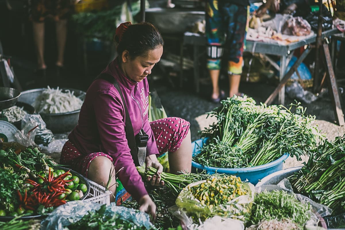 Een vrouw in een roze shirt met lange mouwen en een korte broek zit bij een kraampje op de markt en sorteert bladgroen - een typisch gezicht bij een streetfoodkar in Azië. Grote manden met verse groenten omringen haar, terwijl andere verkopers en producten de drukke achtergrond vullen.