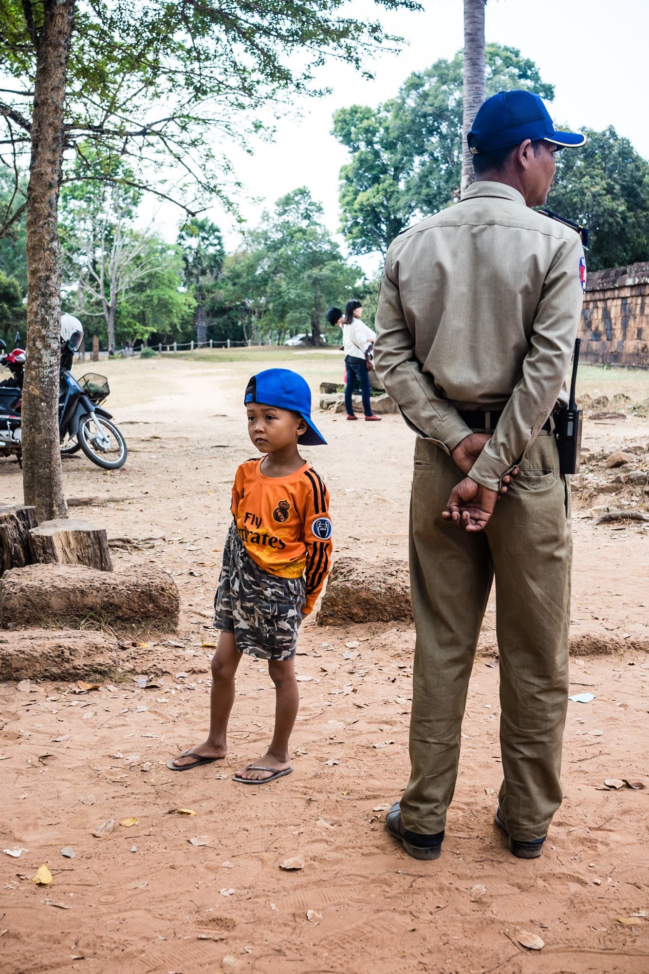 Een jonge jongen met een blauwe pet, een oranje shirt en een camouflageshort staat blootsvoets op een zandpad naast een agent in uniform - een suggestieve scène die de alledaagse portretten van Cambodjanen vastlegt te midden van bomen, motoren en mensen op de achtergrond.