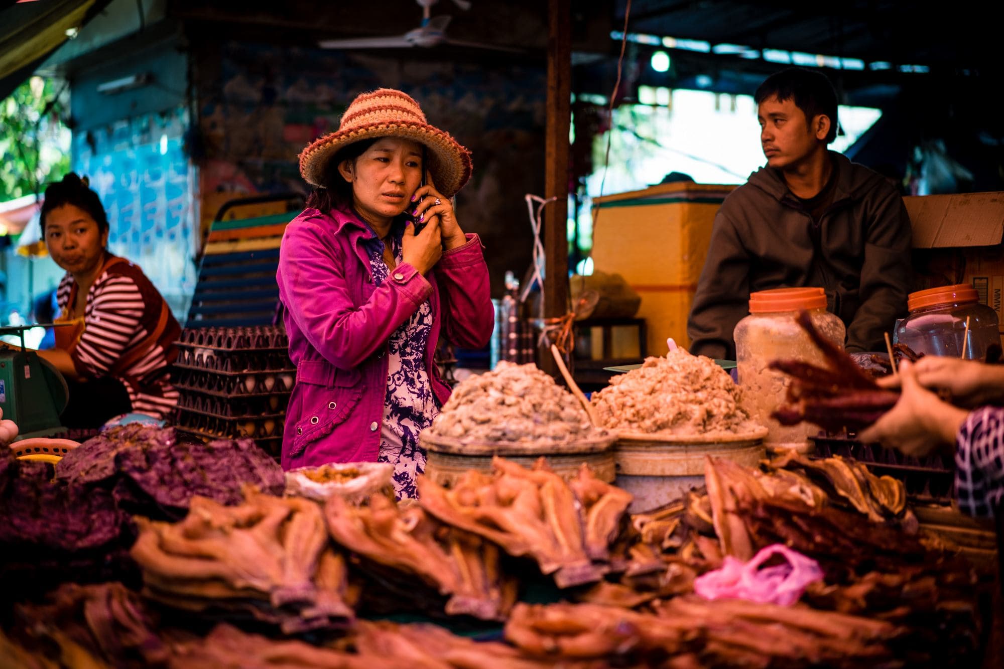 Een vrouw met een roze jas en hoed houdt een telefoon aan haar oor bij een kraampje op de markt, omringd door stapels gedroogd en geconserveerd voedsel - een van de levendige portretten van Cambodjanen te midden van kleurrijke potten en bedrijvigheid.