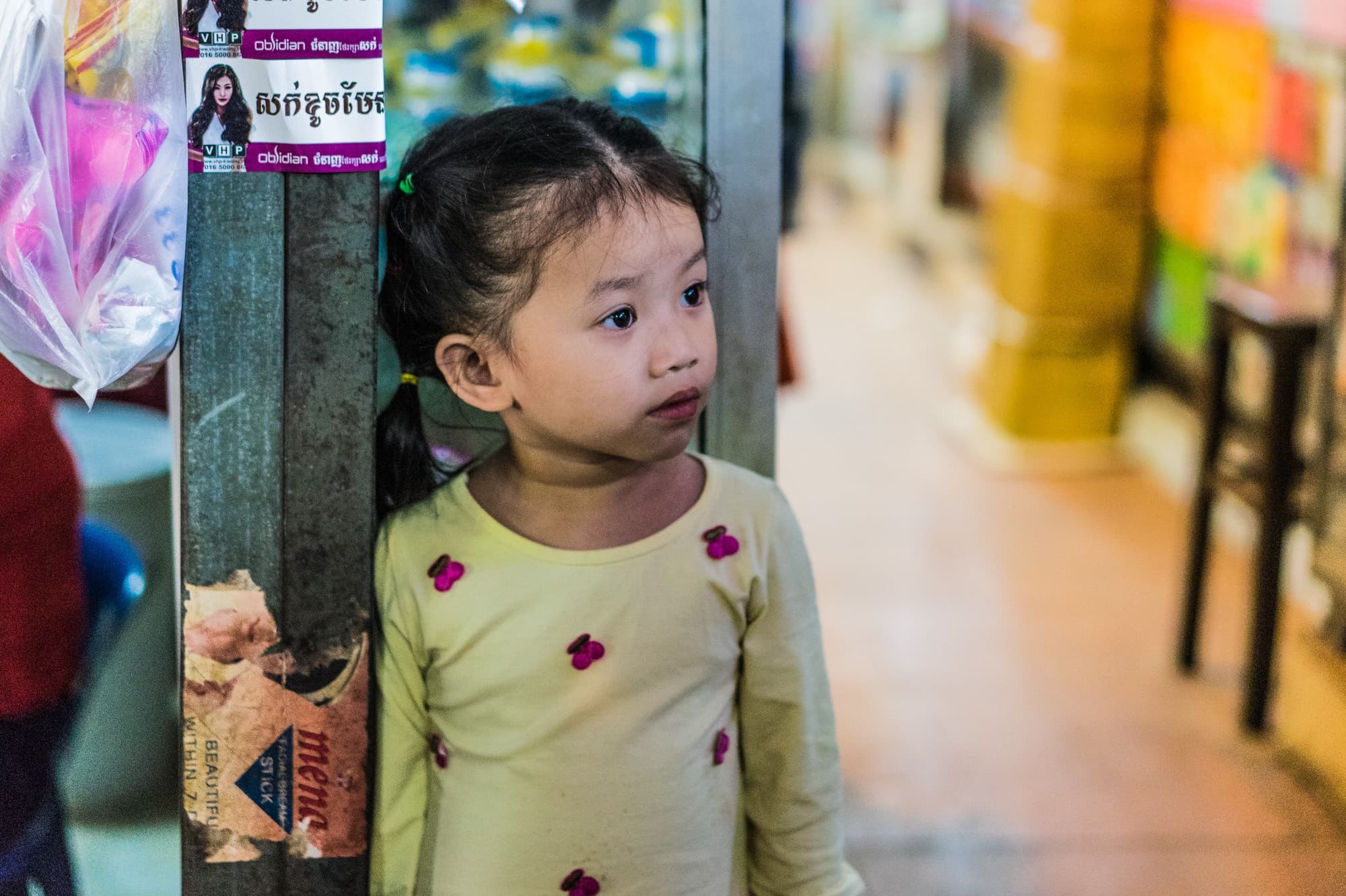 Een jong meisje met gevlochten haar, dat een lichtgeel shirt met kleine roze strikjes draagt, staat binnen naast een metalen paal met Khmer-talige stickers - een levendig voorbeeld van portretten van Cambodjanen op een drukke markt of in een winkel.