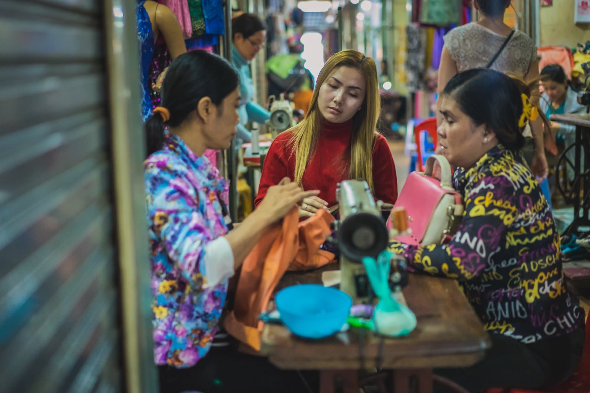 Drie vrouwen zitten aan een tafeltje op een drukke markt. Twee bedienen naaimachines terwijl de derde, in een rode trui, toekijkt. Kleurrijke stoffen hangen om hen heen - een authentiek tafereel onder portretten van Cambodjanen, levendig en geconcentreerd.
