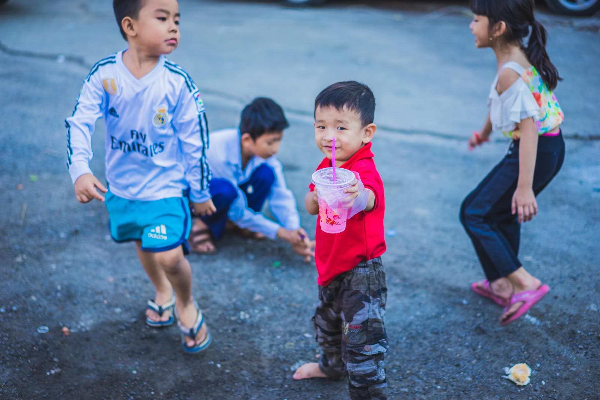 Vier jonge kinderen spelen buiten op een verhard terrein. Eén jongen in een rood shirt en camouflagebroek lacht naar de camera terwijl hij een roze drankje vasthoudt. Dit vrolijke moment legt openhartige portretten van Cambodjanen vast terwijl anderen op de achtergrond spelen.