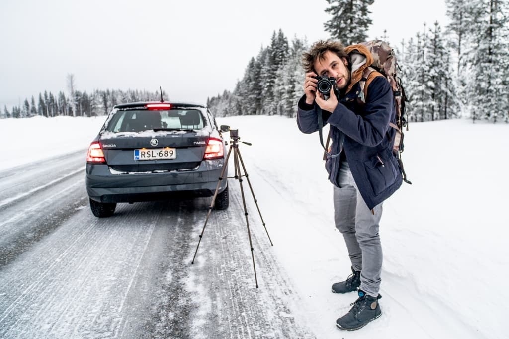 Een persoon met een rugzak staat op een besneeuwde weg naast een zwarte auto en statief, met een camera in de hand om noorderlicht te fotograferen. Besneeuwde bomen vormen de achtergrond onder een bewolkte hemel; bandensporen en voetafdrukken markeren de verse sneeuw.