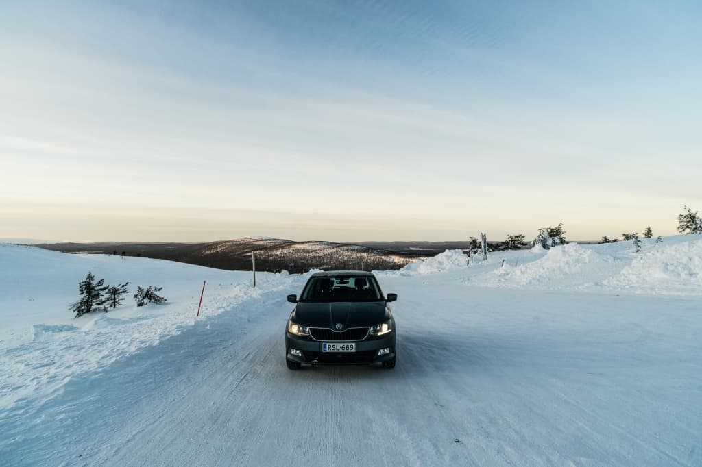 Een zwarte auto staat geparkeerd op een besneeuwde weg omringd door sneeuwbanken en verspreide bomen - een ideale plek voor Noorderlicht fotograferen. Heuvels in de verte en een bleke lucht dragen bij aan het kalme, afgelegen gevoel van dit koude winterlandschap.
