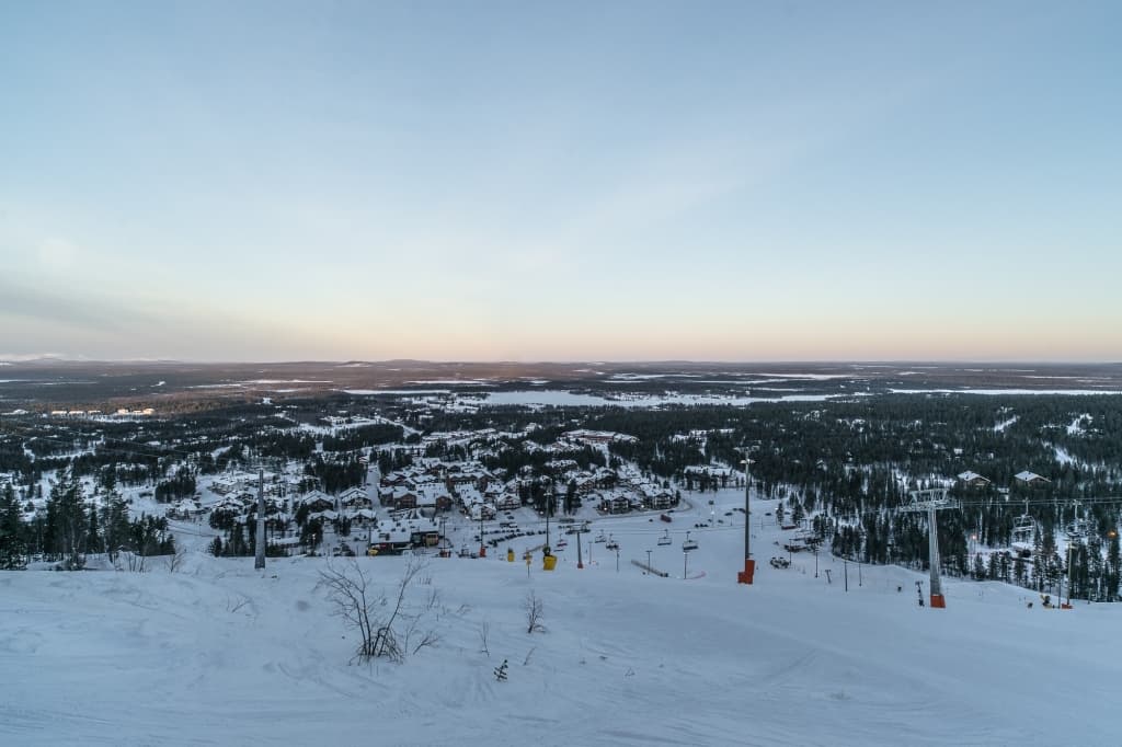Een besneeuwde skipiste kijkt uit over een klein stadje omringd door bossen. Skiliften en skistokken staan langs de helling, terwijl de heldere hemel met een vage gele gloed wijst op een vroege ochtend - perfecte omstandigheden voor noorderlicht fotograferen.