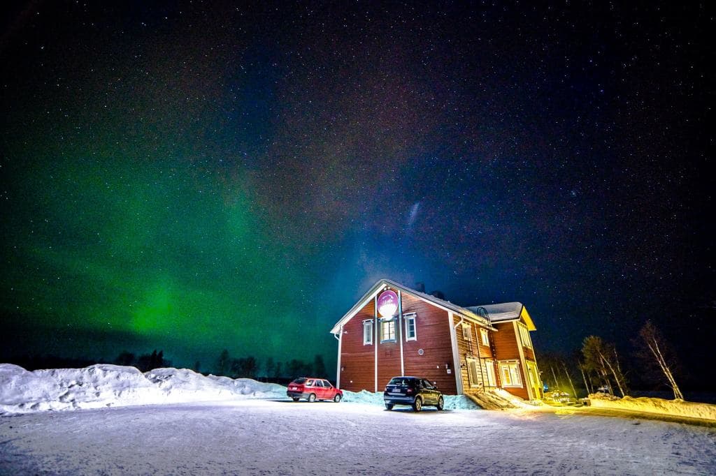 Een besneeuwd landschap bij nacht met een groot houten huis met lichten aan en twee geparkeerde auto's. Daarboven is de nachtelijke hemel gevuld met sterren en een levendig groen noorderlicht, perfect voor noorderlicht fotograferen. Op de achtergrond zijn sneeuwhopen en bomen zichtbaar.