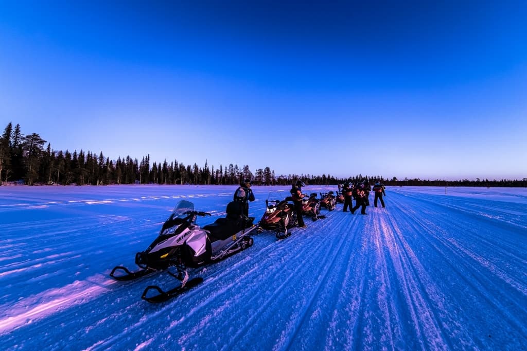Een groep mensen staat naast sneeuwscooters in een rij op een besneeuwd, open landschap in de schemering, klaar om te noorderlicht fotograferen. Schaduwen strekken zich uit over de sneeuw terwijl de diepblauwe lucht vervaagt tot paars achter een rij pijnbomen aan de horizon.