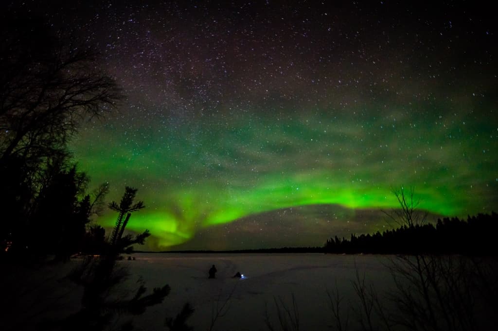 Een nachtelijke hemel toont groene aurora borealis boven een besneeuwd landschap en een bevroren meer, omzoomd door donkere boomsilhouetten. Zwakke sterren zijn zichtbaar terwijl een kleine figuur met een gloeiend licht op het ijs staat en de scène vastlegt terwijl hij noorderlicht fotografeert.