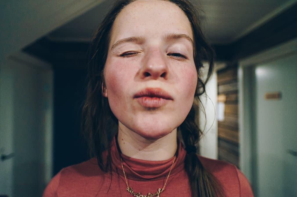 Een jonge vrouw met een lichte huid en sproeten kijkt naar de camera, ogen gedeeltelijk gesloten, lippen samengetrokken alsof ze een kus uitblaast. Ze draagt een mauve coltrui en halsketting, haar losse vlecht zacht verlicht als een noorderlicht gefotografeerd in een hal met witte deuren.