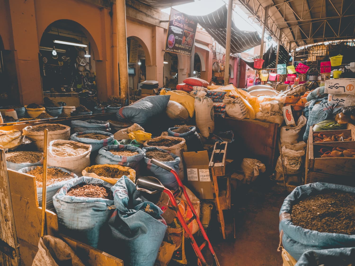 Een bruisend markttafereel dat doet denken aan de souks van Marokko, met zakken en tassen vol specerijen en granen. Kraampjes staan bij elkaar onder een hoog dak, terwijl warm licht naar binnen valt dat kleurrijke uitstallingen verlicht en een levendige, levendige sfeer creëert.