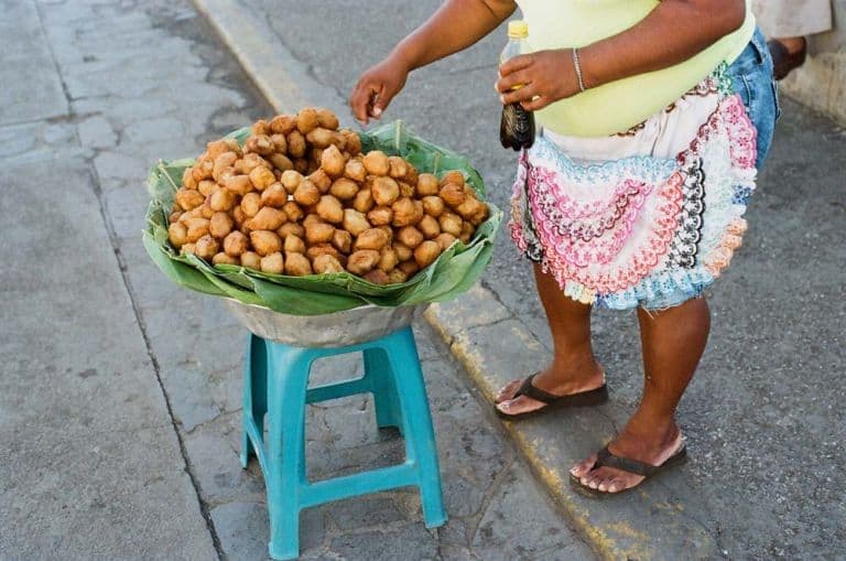 Een persoon met een geel shirt, een kleurrijk schort en slippers staat bij een blauw krukje in Nicaragua, met een metalen schaal bekleed met bananenbladeren en gevuld met ronde, goudbruine gefrituurde hapjes op gebarsten stoeptegels.