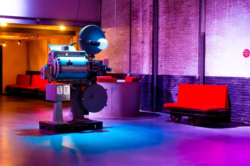 Een vintage filmprojector staat op een gladde, kleurrijk verlichte vloer in een industriële Den Bosch ruimte met bakstenen muren. Rode banken staan langs de muur bij een ronde toonbank en de paarse, blauwe en gele verlichting creëert een op Uitboeren geïnspireerde sfeer.