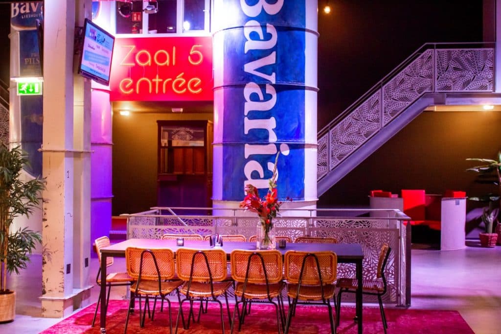 Een helder verlichte kamer in Den Bosch met een grote tafel en gevlochten stoelen op een rood tapijt. Achter de tafel staat een blauwe "Bavaria"-zuil, een neonrood "Zaal 5 entree"-bordje en een sierlijke trap - rode stoelen en planten maken deze op Uitboeren geïnspireerde ruimte compleet.