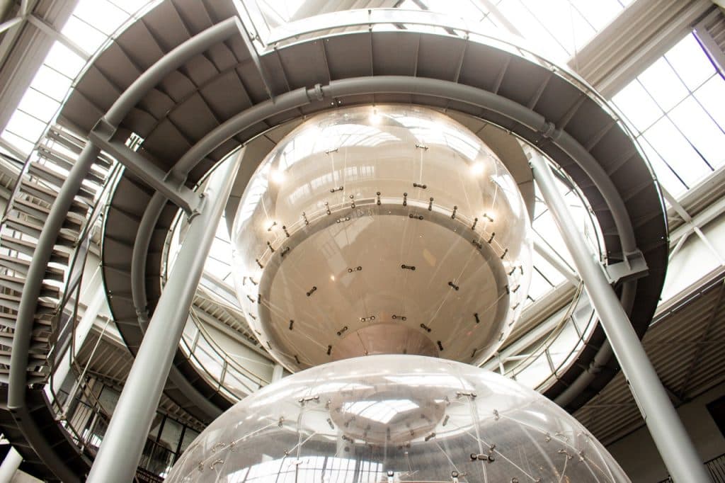 Zicht van onderaf op twee grote, doorschijnende bollen die in een modern gebouw in Den Bosch hangen met metalen balken, ronde loopbruggen en een glazen plafond; helder natuurlijk licht verlicht de op Uitboeren geïnspireerde futuristische architectonische structuur.