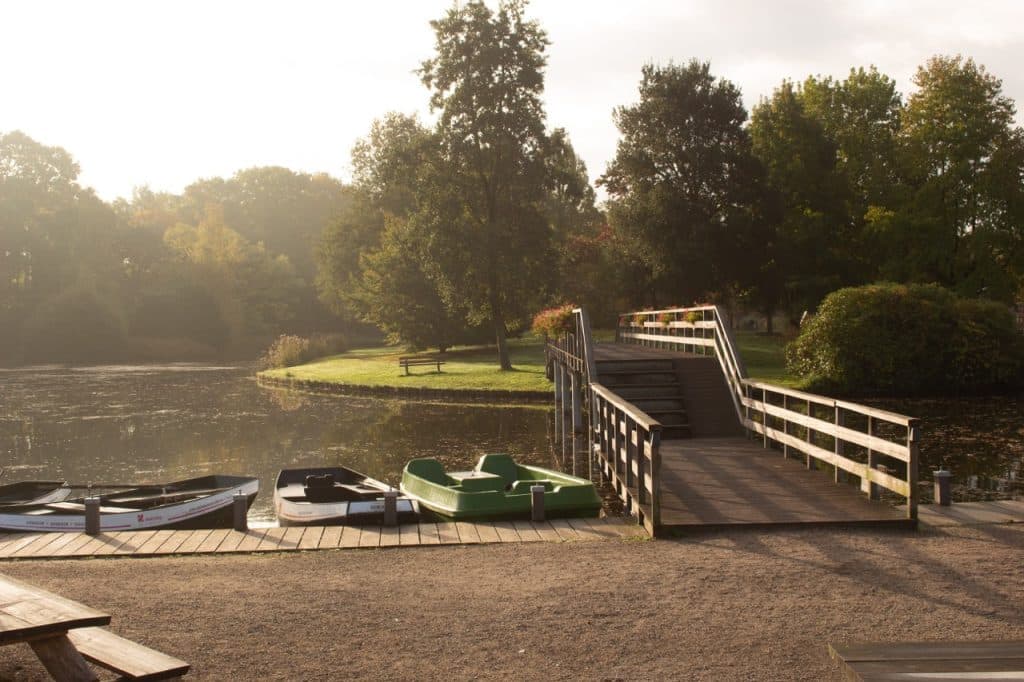 Een vredig tafereel aan het meer bij Den Bosch, met kleine bootjes aangemeerd aan een houten steiger. Een houten helling en brug reiken over het water naar een grasveld met een bankje, omringd door bomen en struiken onder zacht, warm zonlicht - een echt uitboerenverblijf.