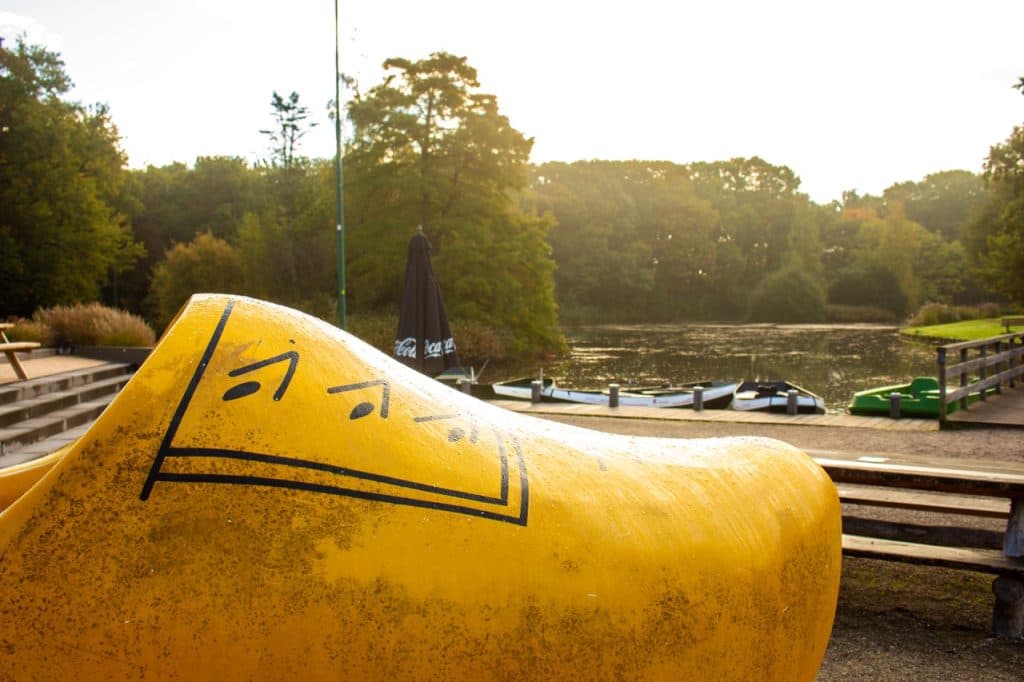 Een grote gele houten klompsculptuur met zwarte markeringen staat bij een rustig meertje omringd door groene bomen in Den Bosch. Verschillende kleine boten liggen aangemeerd op het water en er is een houten pier en bankjes in de buurt onder zacht, gouden zonlicht.