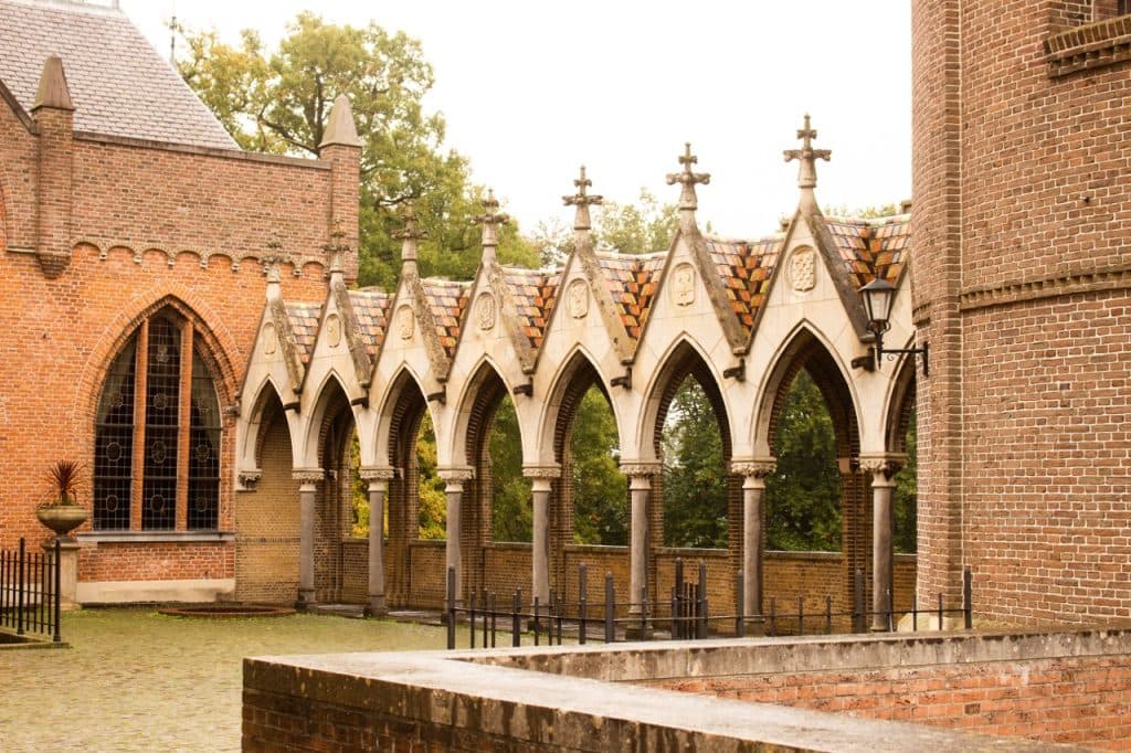 Een rij gotische stenen bogen met spitse toppen, kruisbloemstukken en pannendaken verbindt twee bakstenen gebouwen in Den Bosch. Grote boogramen, ijzeren hekken en groene bomen verschijnen op de achtergrond onder een bewolkte hemel.