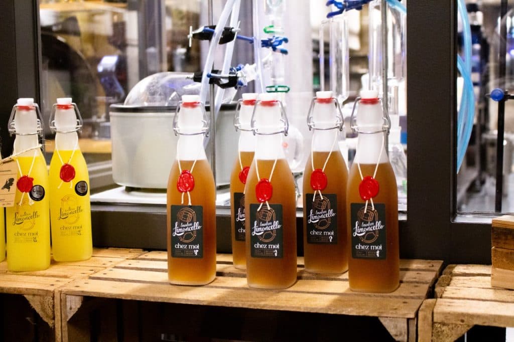 Glazen flessen gevuld met amberkleurige en gele vloeistoffen staan op houten kratten in een ambachtelijke drankensetting die doet denken aan Uitboeren in Den Bosch. De amberkleurige flessen zijn verzegeld met rode was, terwijl de gele flessen voorzien zijn van bleke labels te midden van laboratoriumapparatuur en pijpen.