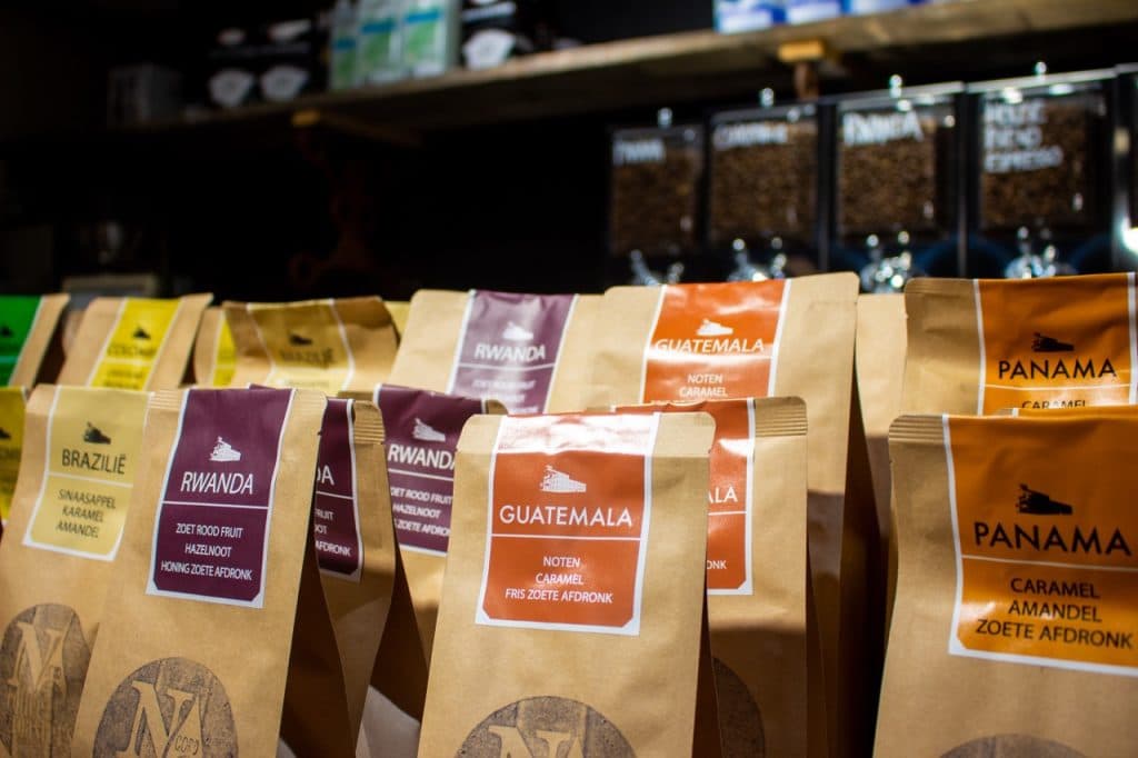 Zakken speciale koffiebonen uit Brazilië, Rwanda, Guatemala en Panama staan uitgestald op een plank in Den Bosch. Elke zak is voorzien van een kleurrijk etiket met landnamen en proefnotities. Uitboeren koffiebonenautomaten zijn zichtbaar in de onscherpe achtergrond.