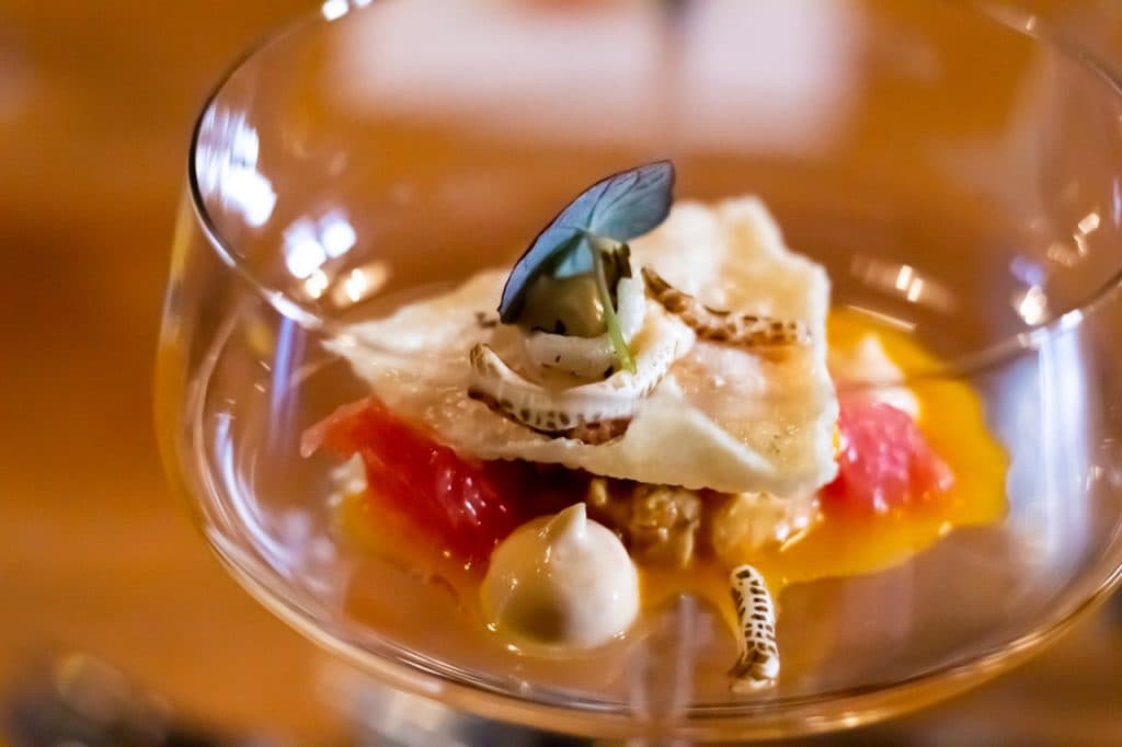 Een heldere glazen schaal toont een gastronomisch voorgerecht met een blauwe vlindervleugel, room, een cracker, roze grapefruit, saus en eetbare insecten - een kunstzinnige mix van texturen en kleuren geïnspireerd door de innovatieve culinaire scene in Den Bosch.