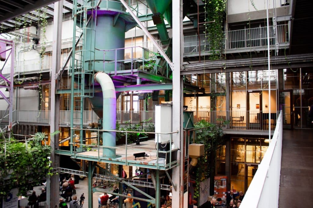 Een moderne binnenruimte in Den Bosch met zichtbare metalen constructies, buizen en groene planten die tegen de muren klimmen. Grote industriële leidingen en kantoren met glazen wanden kijken uit op een gemeenschappelijke ruimte waar mensen samenkomen - een sfeer die echt past bij Uitboeren.