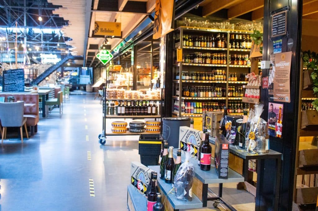 Moderne overdekte markt in Den Bosch met schappen met drank in flessen, cadeaumanden en levensmiddelen. Aan de linkerkant staan eettafels; rechts staan bieren en snacks van Uitboeren. Heldere verlichting en borden aan het plafond creëren een uitnodigende sfeer.