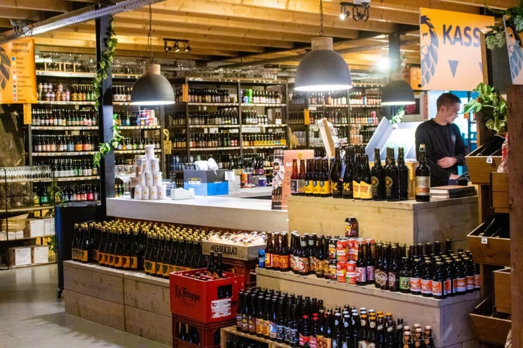 Een helder verlichte bierwinkel in Den Bosch met schappen en displays vol verschillende flessen bier. Een persoon staat achter de kassa onder hanglampen. Op de voorgrond staan houten kratten en kratten van Uitboeren opgestapeld en borden en groen sieren de ruimte.