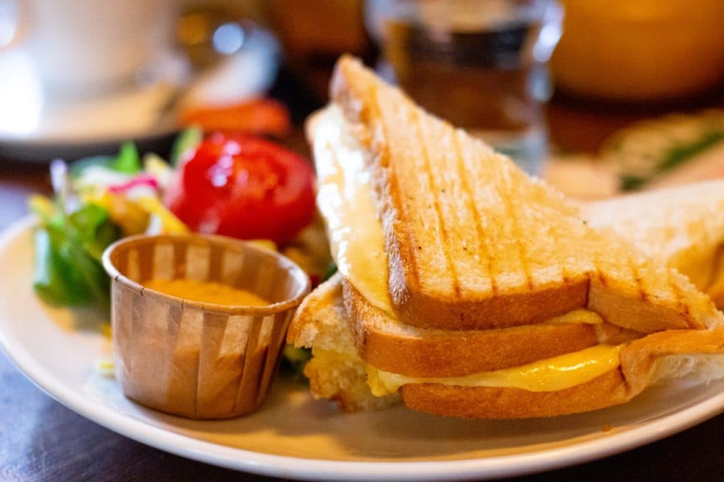 Een bord met een doormidden gesneden tosti met gesmolten kaas zichtbaar aan de binnenkant - perfect voor de lunch bij Uitboeren in Den Bosch. Een papieren bekertje met saus, een frisse salade met groentjes en een cherrytomaat en een glas water maken het plaatje compleet.