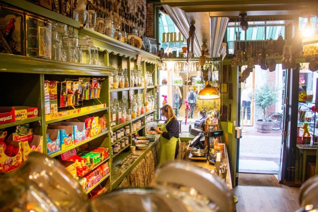 Een vrouw staat achter de toonbank in een gezellige snoepwinkel in vintage-stijl in Den Bosch met potten, planken met kleurrijk snoepgoed en een nostalgische inrichting. Warme verlichting vult de ruimte en zonlicht stroomt door de open deur naar binnen naar mensen buiten op straat.