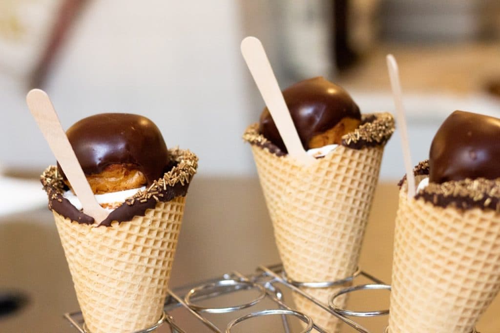 Drie wafelhoorntjes gedoopt in chocolade en bestrooid met chocoladeschaafsel zijn gevuld met room en belegd met chocoladegebak, elk met een houten lepel erin, rechtop uitgestald in een metalen rek bij Uitboeren in Den Bosch.