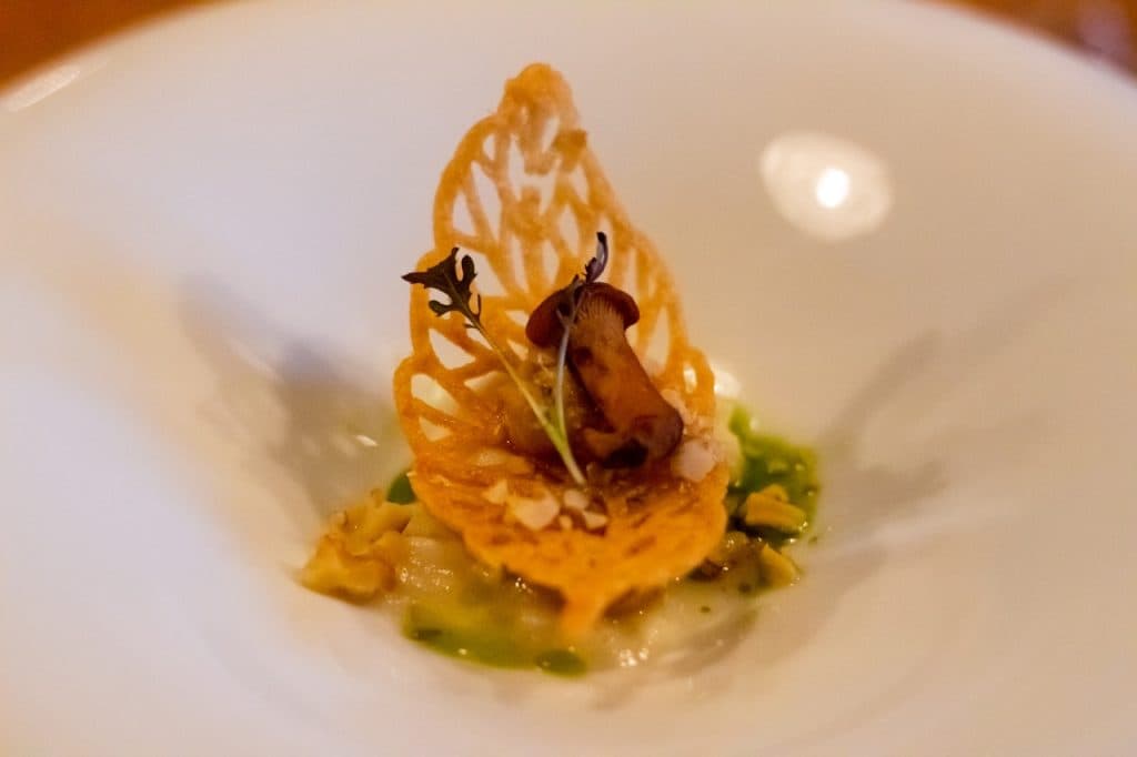 Op een delicaat bord bij Uitboeren in Den Bosch staat een kanten, goudbruine krokant rechtop, met daarop een kleine champignon en microgroenten, rustend op een romige saus met groene kruidenolie en fijngehakte garnering.